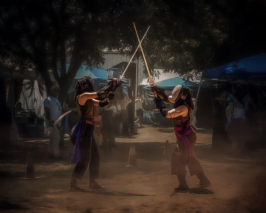 Two performers in Renaissance-inspired costumes sword fighting beneath the trees at the West Texas Renaissance Festival in Odessa, Texas.